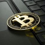 Złota fizyczna moneta Bitcoin jest umieszczona na klawiaturze laptopa. Logo Bitcoin, duże „B” z dwiema liniami, jest wyraźnie widoczne w środku monety. Klawisze laptopa i touchpad są widoczne w tle.