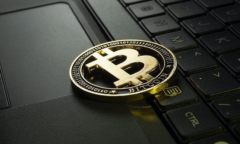 Złota fizyczna moneta Bitcoin jest umieszczona na klawiaturze laptopa. Logo Bitcoin, duże „B” z dwiema liniami, jest wyraźnie widoczne w środku monety. Klawisze laptopa i touchpad są widoczne w tle.