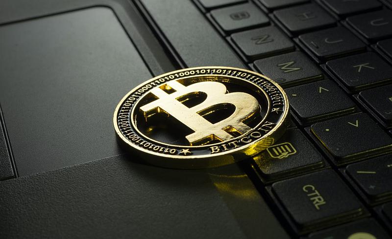 Złota fizyczna moneta Bitcoin jest umieszczona na klawiaturze laptopa. Logo Bitcoin, duże „B” z dwiema liniami, jest wyraźnie widoczne w środku monety. Klawisze laptopa i touchpad są widoczne w tle.