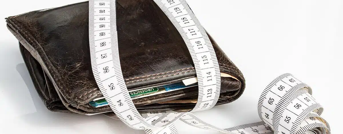 Zużyty skórzany portfel wypełniony kartami i gotówką jest owinięty białą taśmą mierniczą, co sugeruje ograniczenia finansowe lub budżetowanie. Tło jest białe.