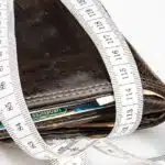 Zużyty skórzany portfel wypełniony kartami i gotówką jest owinięty białą taśmą mierniczą, co sugeruje ograniczenia finansowe lub budżetowanie. Tło jest białe.